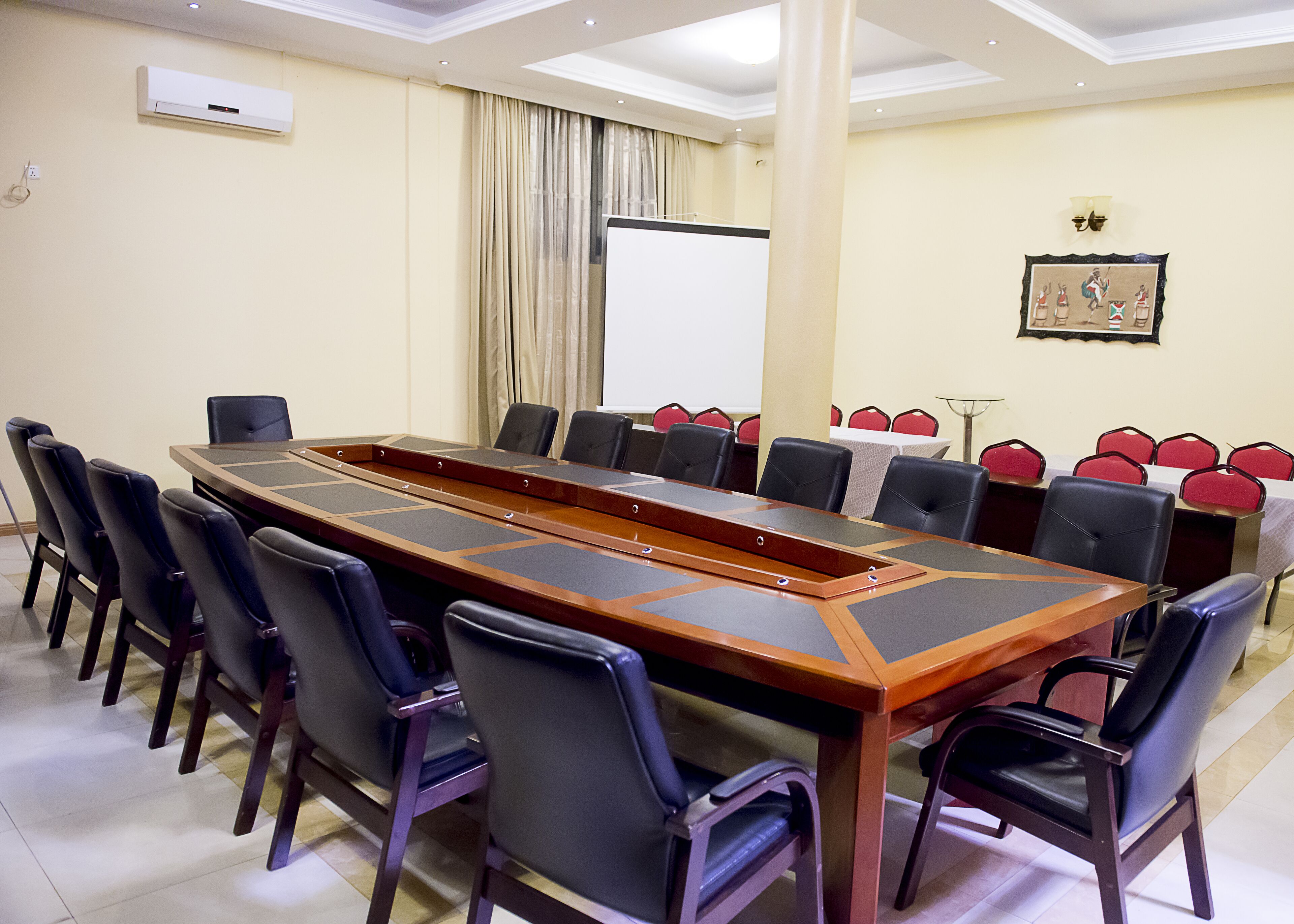 Un salón de conferencias con una larga mesa, varias sillas, una pantalla de proyector y un cuadro en la pared.