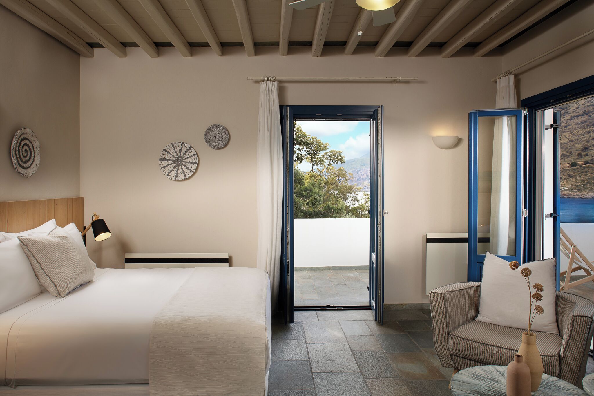 A bedroom with a bed, a chair, a small table with a vase, a window with curtains, and a view of the outdoors.