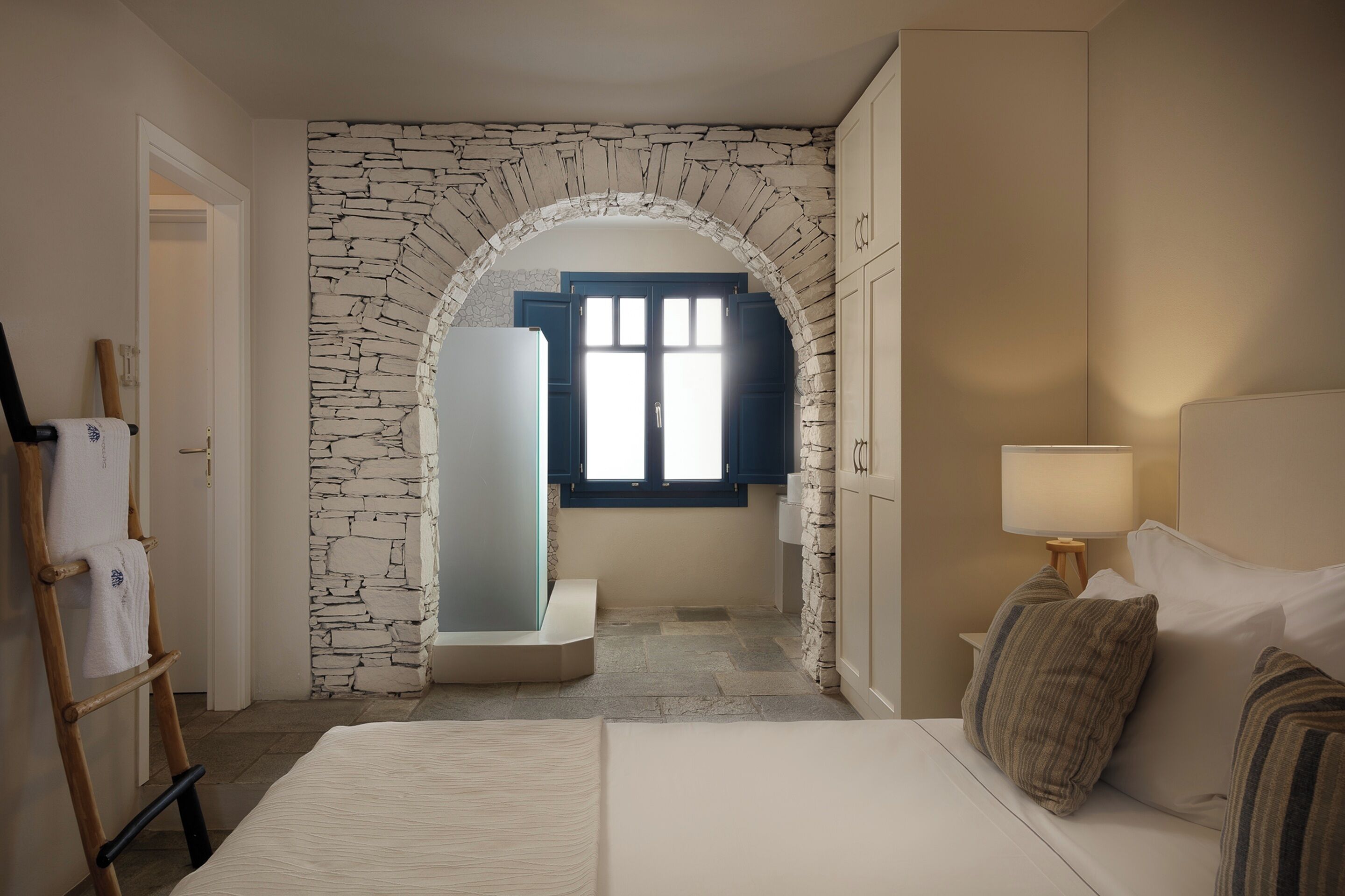 Ein Schlafzimmer mit einem Steingewölbe, einem Bett, einem Nachttisch, einer Lampe und Blick durch ein Fenster.