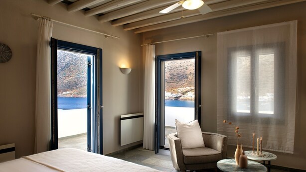 A hotel room with a bed, a chair, a small table with candles, and a view of the sea through open doors. - Sifnos House - Rooms & Spa (Sifnos)