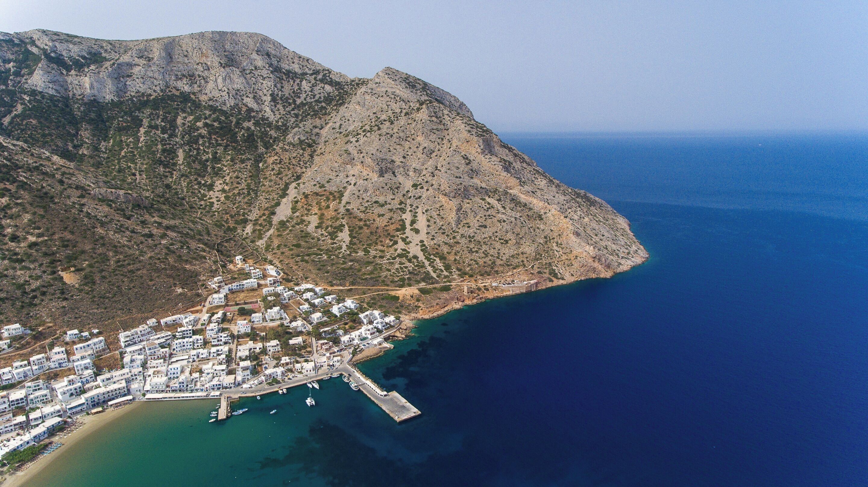 An aerial view of a coastal town with white buildings, a marina, and a mountainous backdrop.