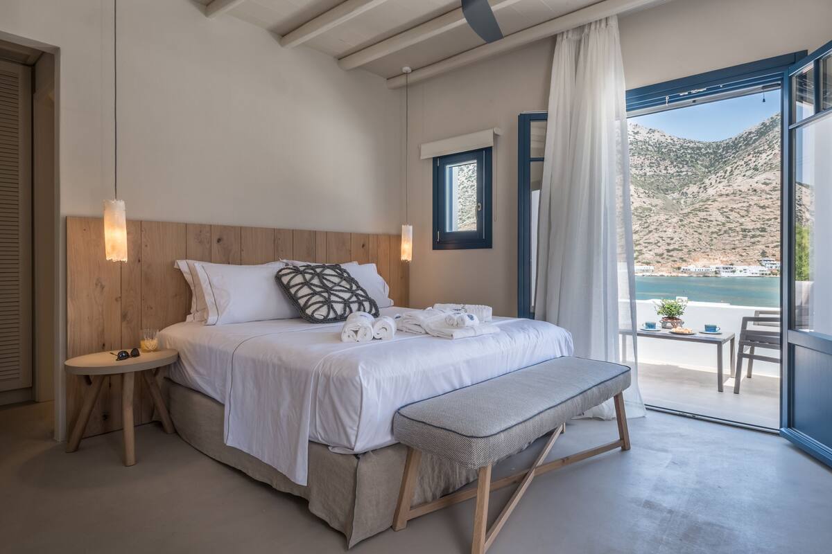 Ein Schlafzimmer mit einem hölzernen Kopfteil, einer Bank, einem Nachttisch mit Lampe und Blick auf das Meer durch ein großes Fenster.