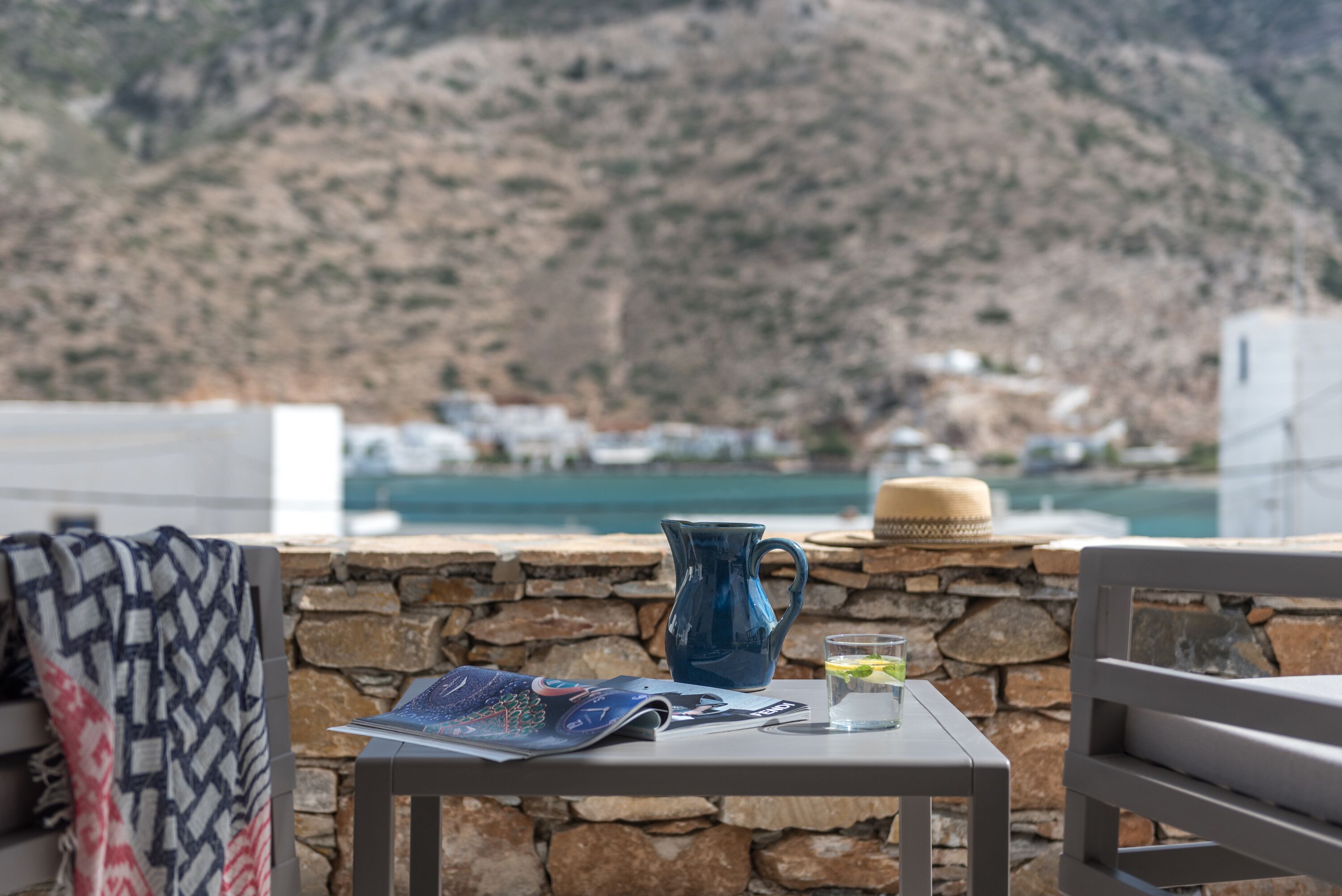Ein Tisch mit einem blauen Krug, einem Glas Wasser und einer Speisekarte auf einer Terrasse mit Blick auf eine gebirgige Landschaft, im Hintergrund weiße Gebäude.