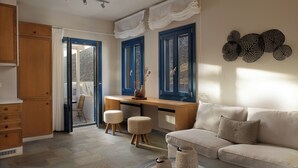 A modern living room with a sofa, a wooden desk, and two ottomans. There is a view through a glass door to an outdoor area. - Sifnos House - Rooms & Spa (Sifnos)