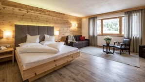 Un dormitorio con cabecera de madera, cama con ropa blanca, mesita de noche, sofá, sillón, una pequeña mesa y una ventana con cortinas.