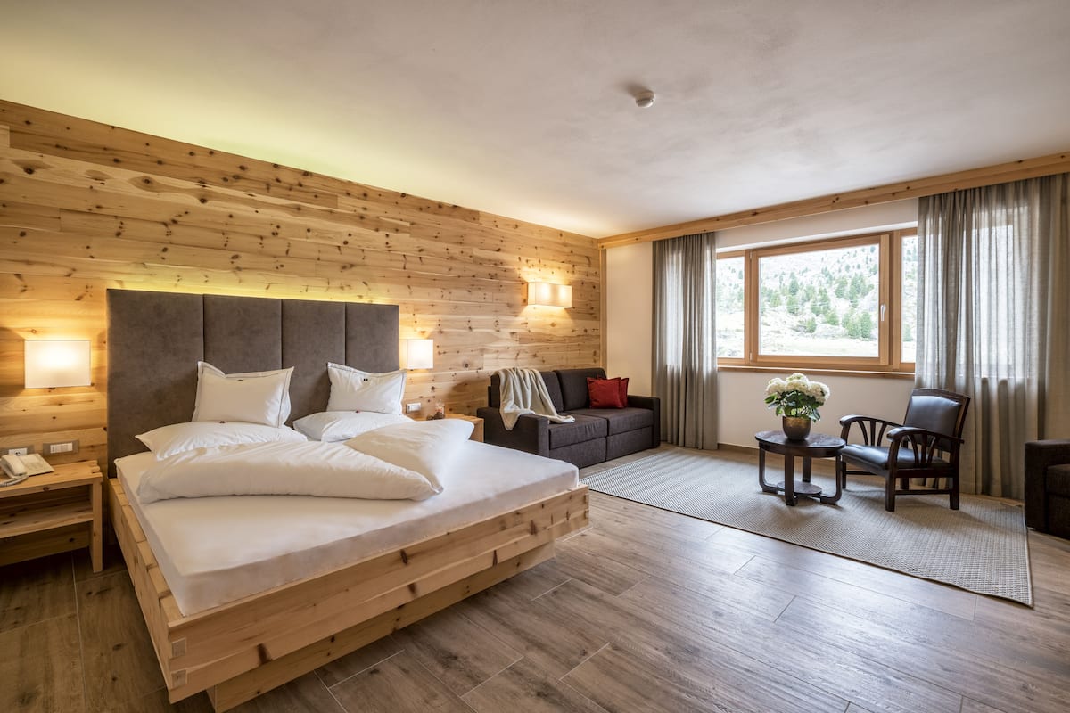 A bedroom with a wooden headboard, a bed with white linens, a nightstand, a sofa, a chair, a small table, and a window with curtains.