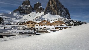 Un albergue cubierto de nieve en la montaña con varios edificios de madera, una pista de esquí en primer plano y un imponente pico rocoso al fondo.