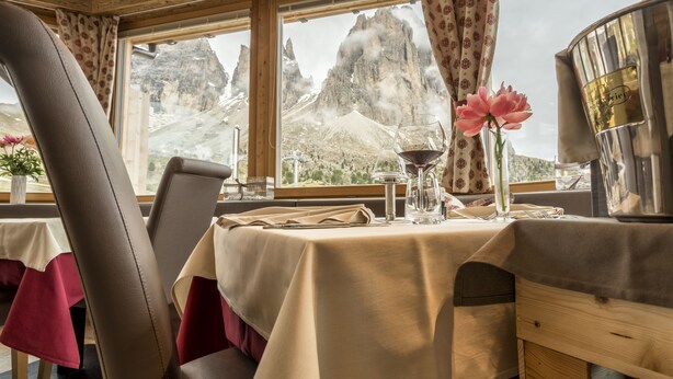 Una mesa de comedor dispuesta con una copa de vino, un florero con una flor y un recipiente metálico, con vistas a la montaña a través de la ventana.