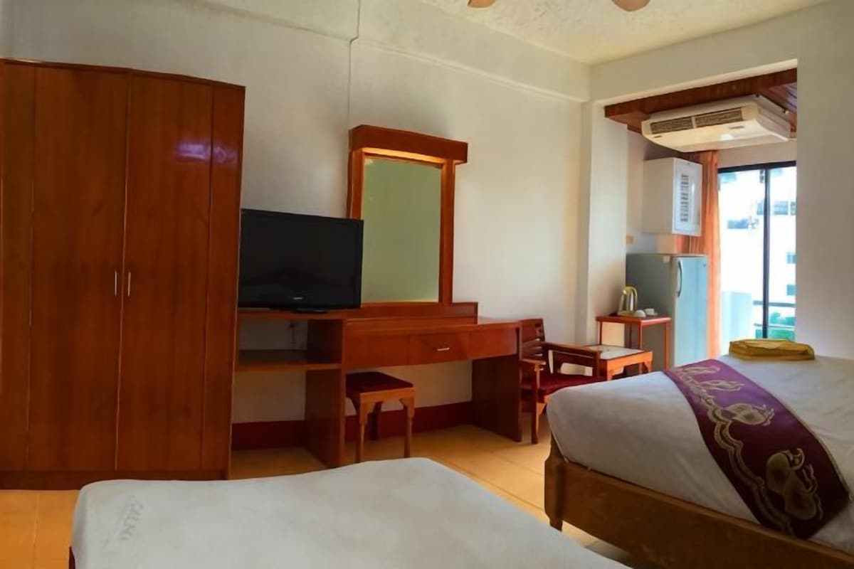 A hotel room with a bed, a desk with a television, a wardrobe, and a window with an air conditioning unit.