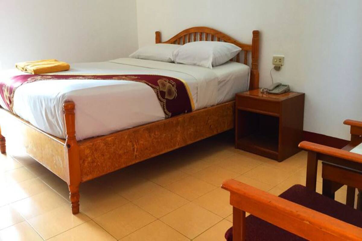 A wooden bed with a white and maroon bedspread, a nightstand with a telephone, and a wooden chair.