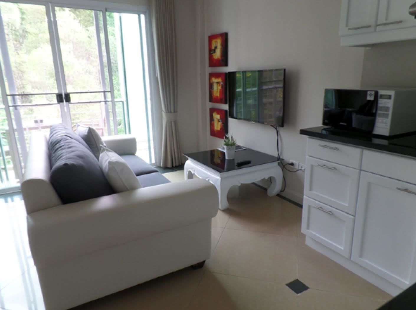 A modern living room with a sofa, a coffee table, and a TV. There is a kitchen area with cabinets and a microwave.