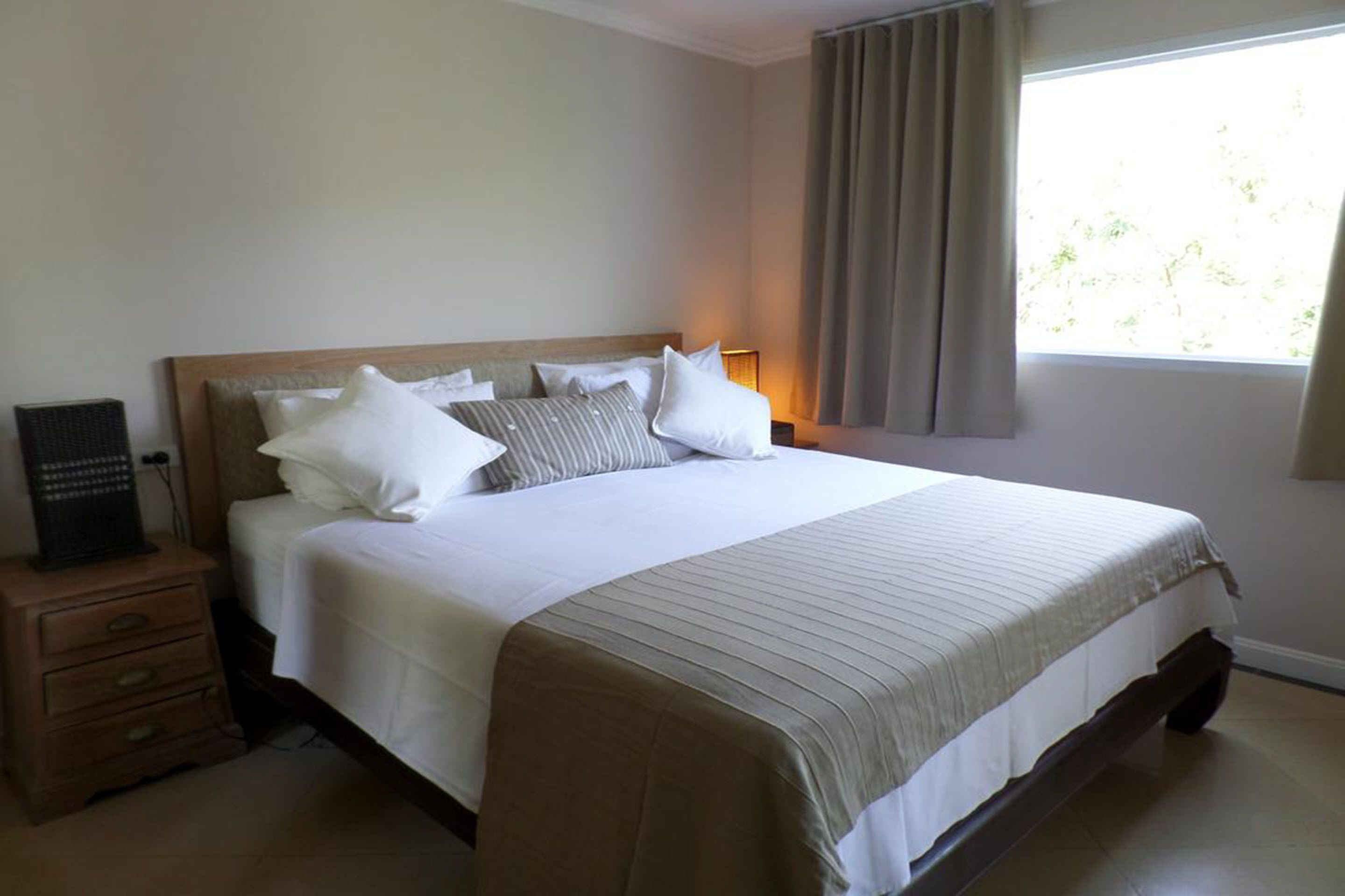 Una cama bien hecha con sábanas blancas y varias almohadas, una mesita de noche con lámpara y una ventana con cortinas.