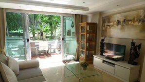 A modern living room with a glass coffee table, a sofa, a TV on a white cabinet, and a sliding glass door leading to an outdoor area. - Westbury Residence (Pattaya)