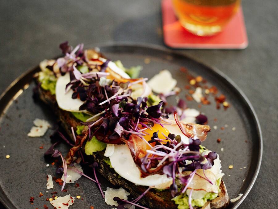 A plate with a sandwich topped with greens, egg, and cheese, and a glass of beer in the background.