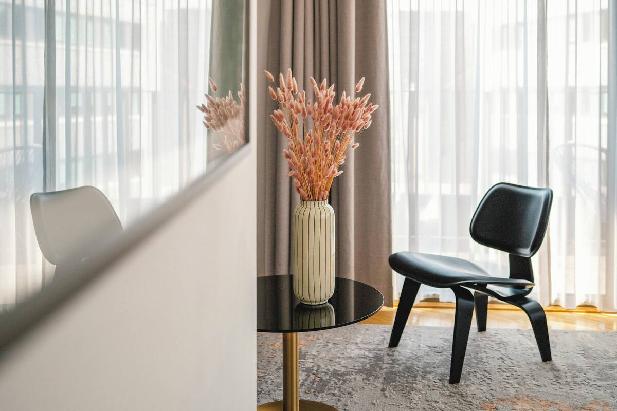 Une chambre d’hôtel moderne avec une grande fenêtre, une table ronde ornée d’un vase de fleurs séchées, une chaise noire et une moquette à motifs.