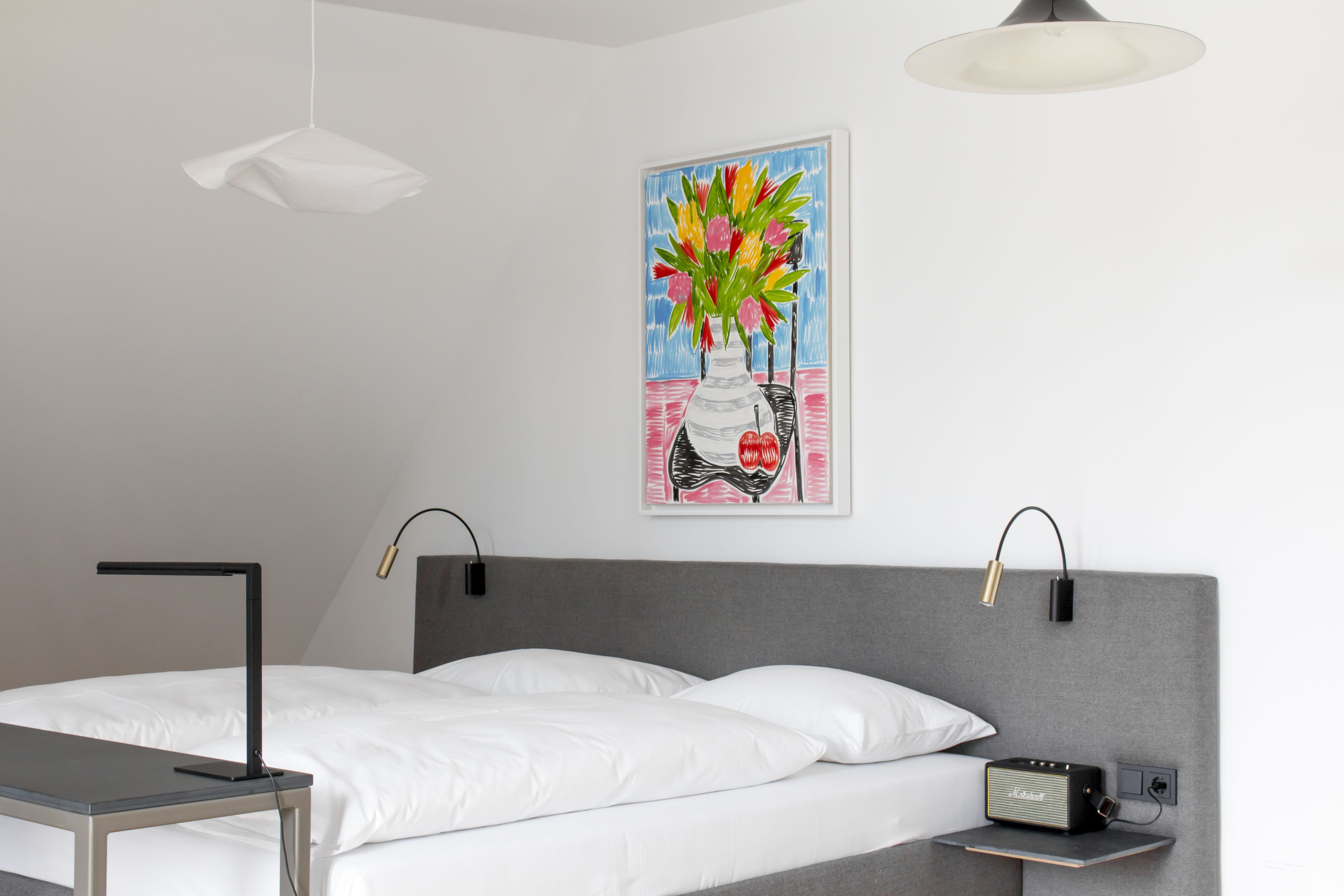 A modern hotel room with a bed, bedside table, lamp, and a painting of a vase with flowers on the wall.
