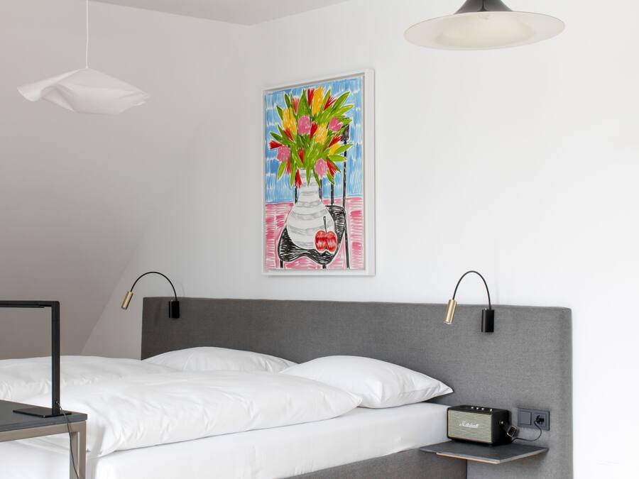 A modern hotel room with a bed, bedside table, lamp, and a painting of a vase with flowers on the wall.