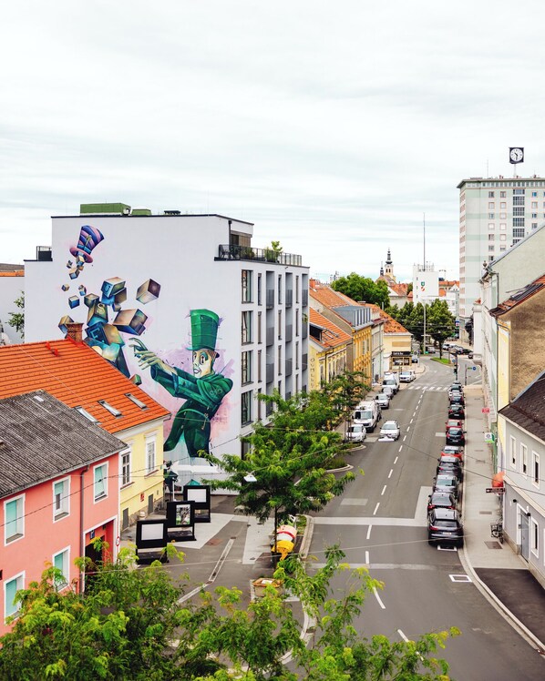 A street view with a large mural on a building, cars parked along the street, and various buildings in the background.