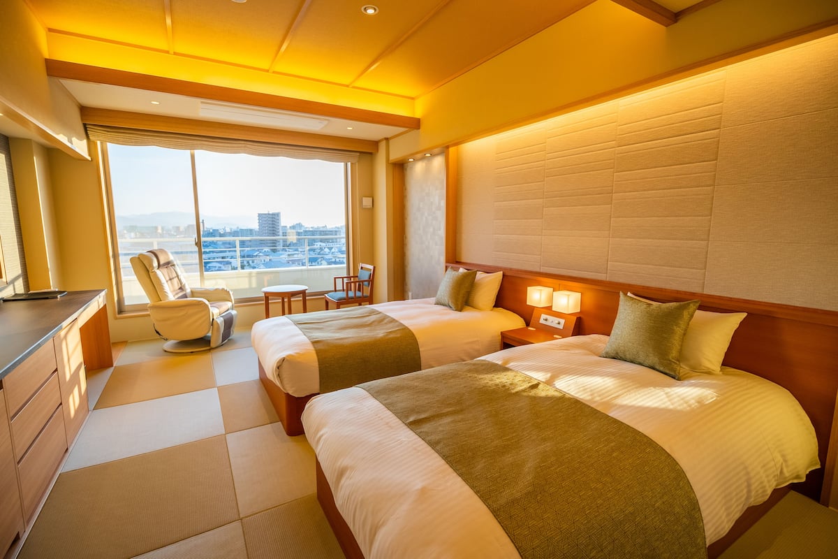 A hotel room with two beds, a desk, and a chair. There is a large window with a city view.