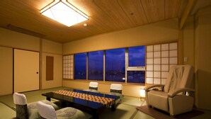 A traditional Japanese room with tatami flooring, a low table with a woven mat, a cushioned seating area, and a view of the city at night through large windows. - Dogo Prince Hotel (Matsuyama)