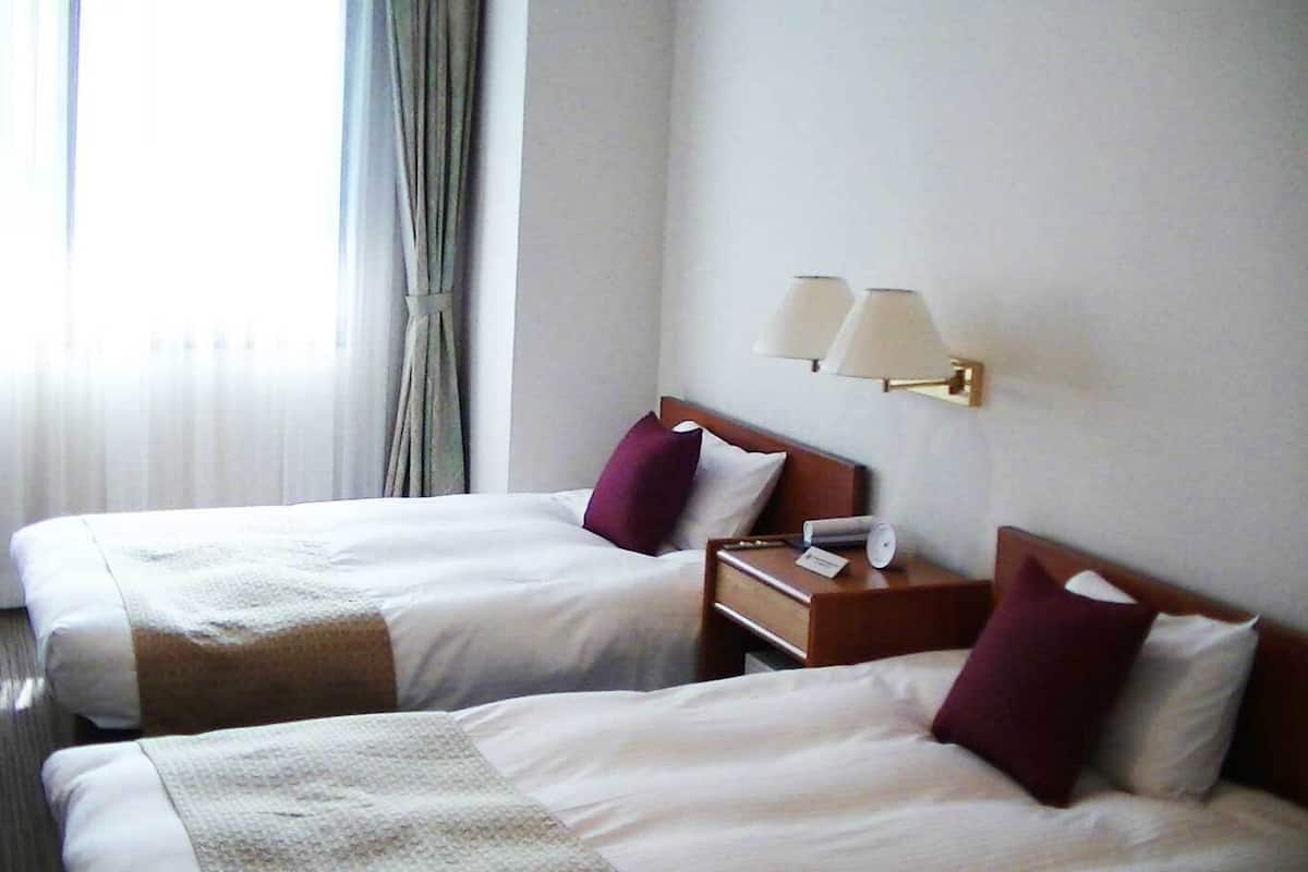 A hotel room with two beds, a nightstand with a lamp, and a window with curtains.