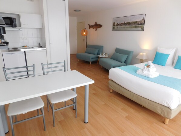 Ett modernt hotellrum med ett litet pentry, en säng med blått sänglinne, ett skrivbord med stolar och ett litet bord med en lampa.