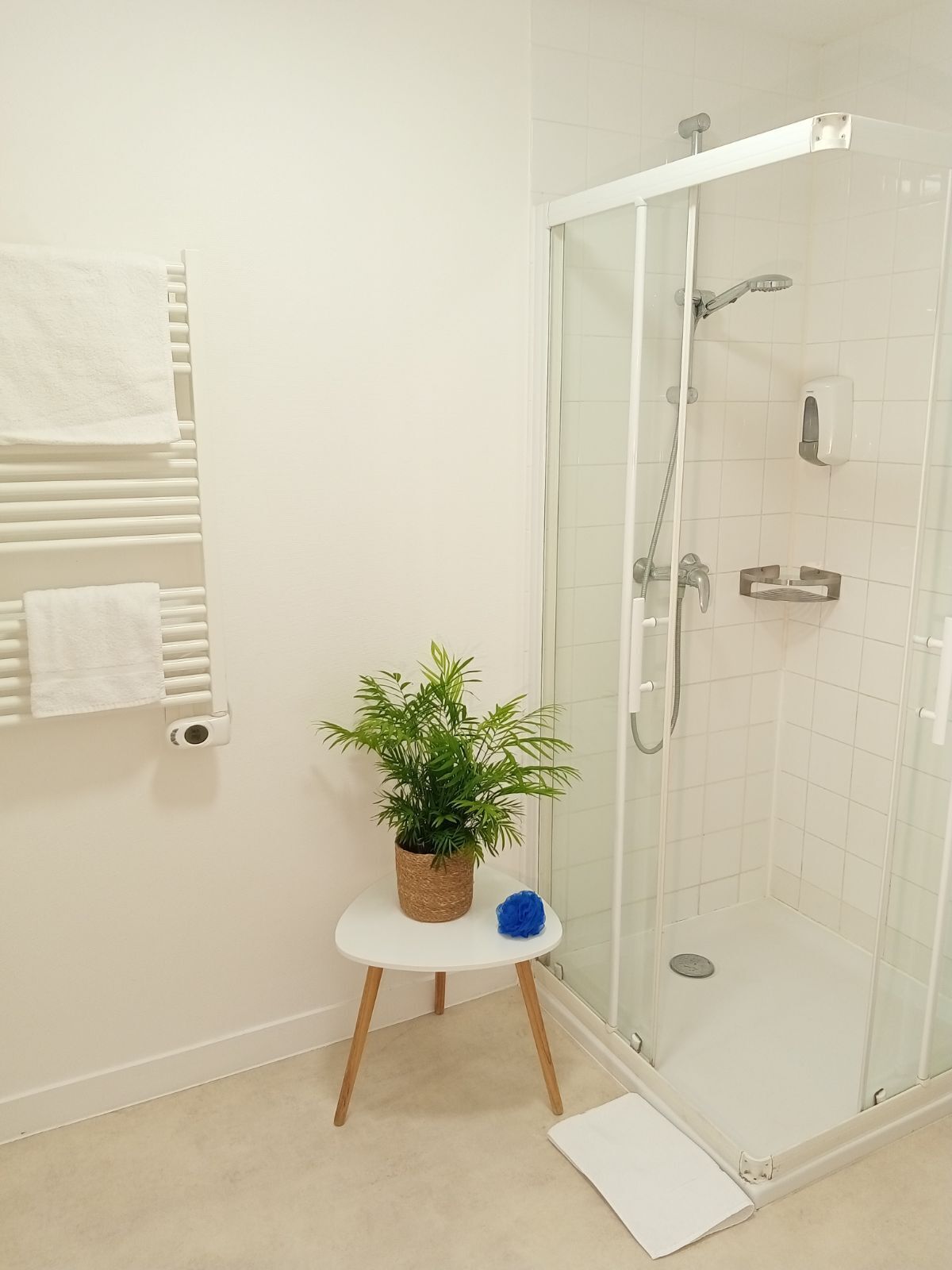 Un baño moderno con cabina de ducha de vidrio, paredes cubiertas de azulejos blancos, un taburete de madera con una planta en maceta y un toallero blanco.