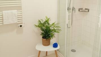 Un baño moderno con cabina de ducha de vidrio, paredes cubiertas de azulejos blancos, un taburete de madera con una planta en maceta y un toallero blanco.