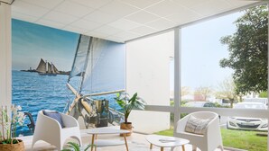 Une pièce dotée d’une grande fenêtre, d’une fresque représentant un voilier, de meubles blancs et offrant une vue sur la circulation et la verdure environnante.
