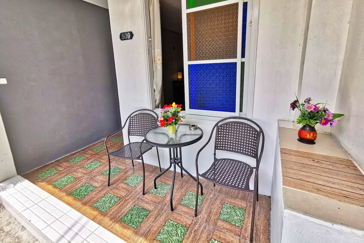 A small outdoor seating area with a table and two chairs, a window with a view, and a door with a number sign.