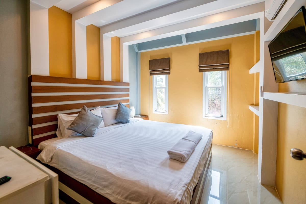 A hotel room with a large bed, a wooden headboard, two windows with blinds, a flat-screen TV mounted on the wall, and a small bedside table.