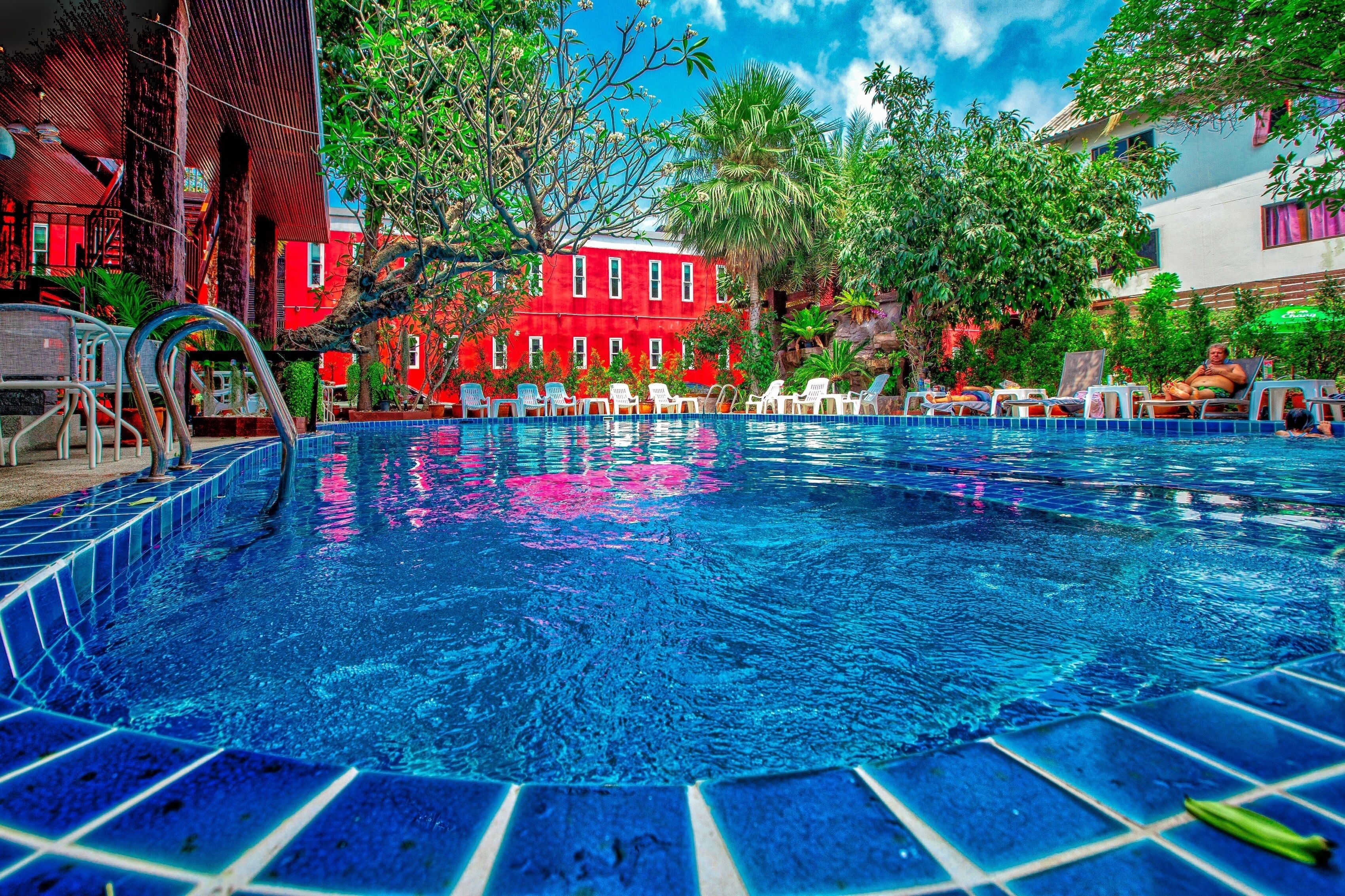 A swimming pool with a tiled edge, surrounded by lounge chairs and a red building in the background.