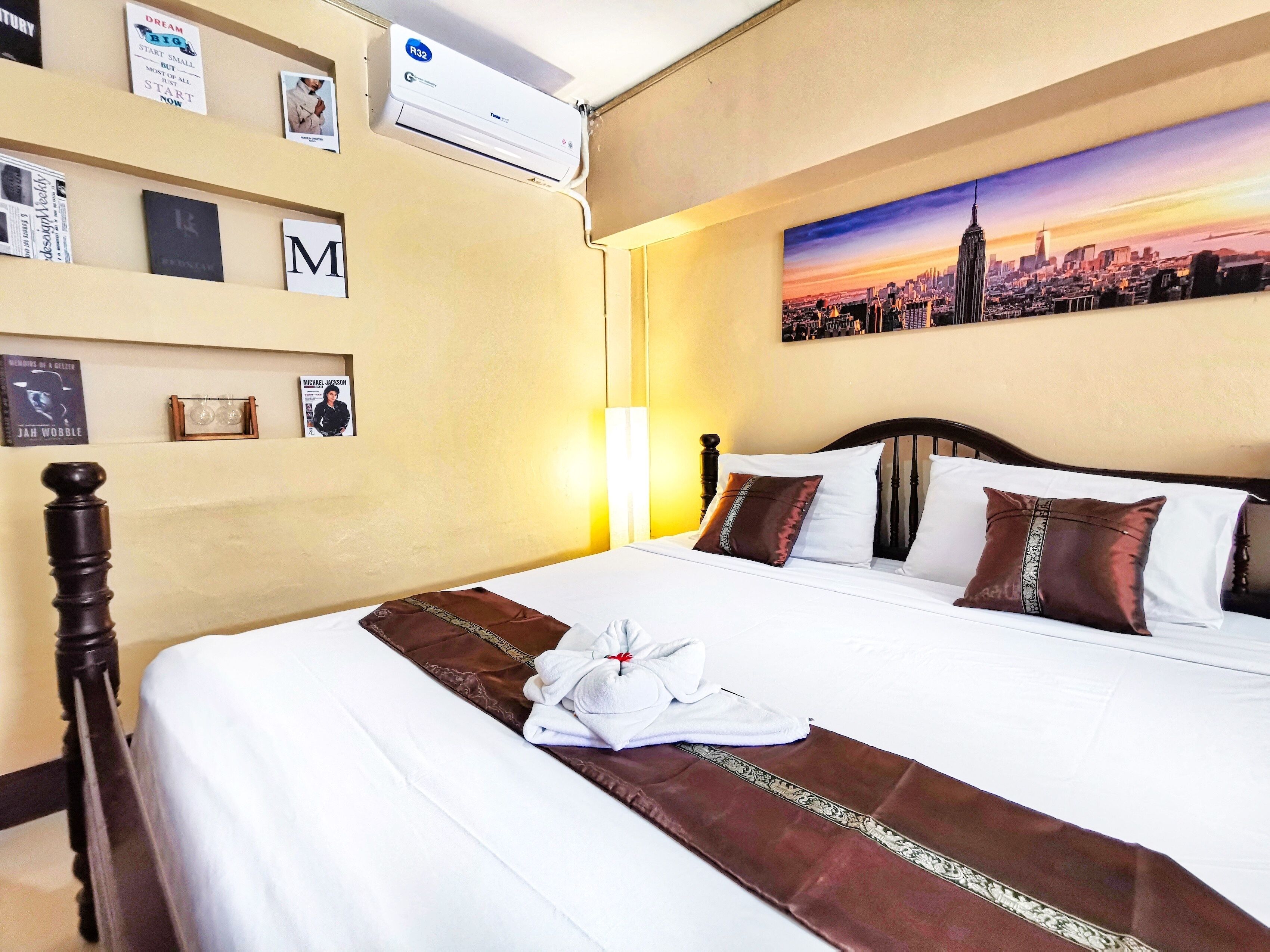 A hotel room with a neatly made bed, a cityscape painting on the wall, and a wall-mounted air conditioner.
