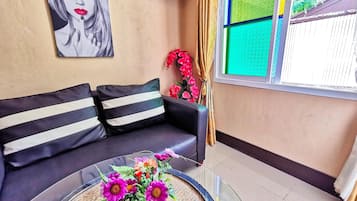 A living room with a black sofa, a glass coffee table with a vase of flowers, a painting on the wall, and a window with patterned curtains.