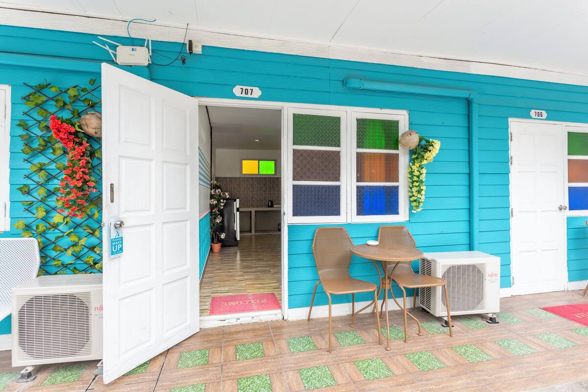 A turquoise building with a white door marked '707' and a red mat. Inside, there are two chairs and a table.