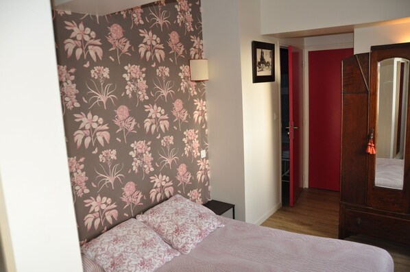 Et soveværelse med blomstret tapet, en seng med pink sengetøj, en sengetårnslampe, en træskab og en rød dør i baggrunden.