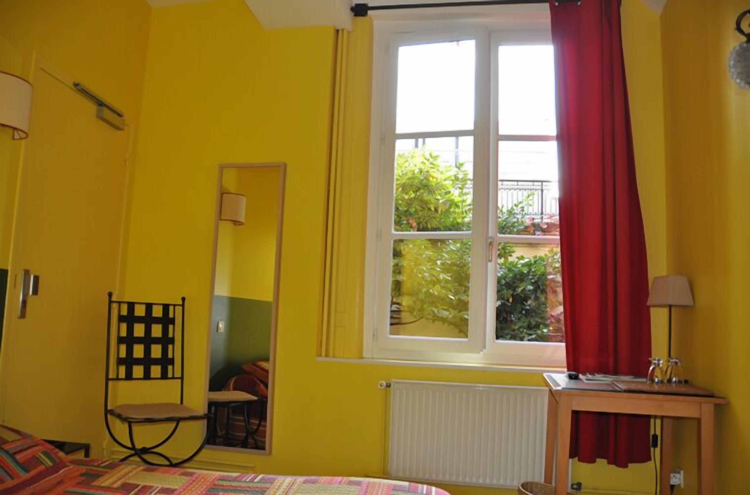 A room with yellow walls, a bed, a chair, a table with a lamp, and a window with red curtains.