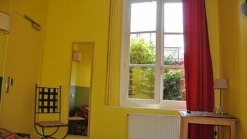 A room with yellow walls, a bed, a chair, a table with a lamp, and a window with red curtains.
