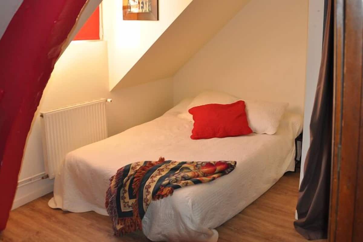 Une petite chambre avec un lit simple, une couvre-lit blanche, un oreiller rouge et une couverture colorée.
