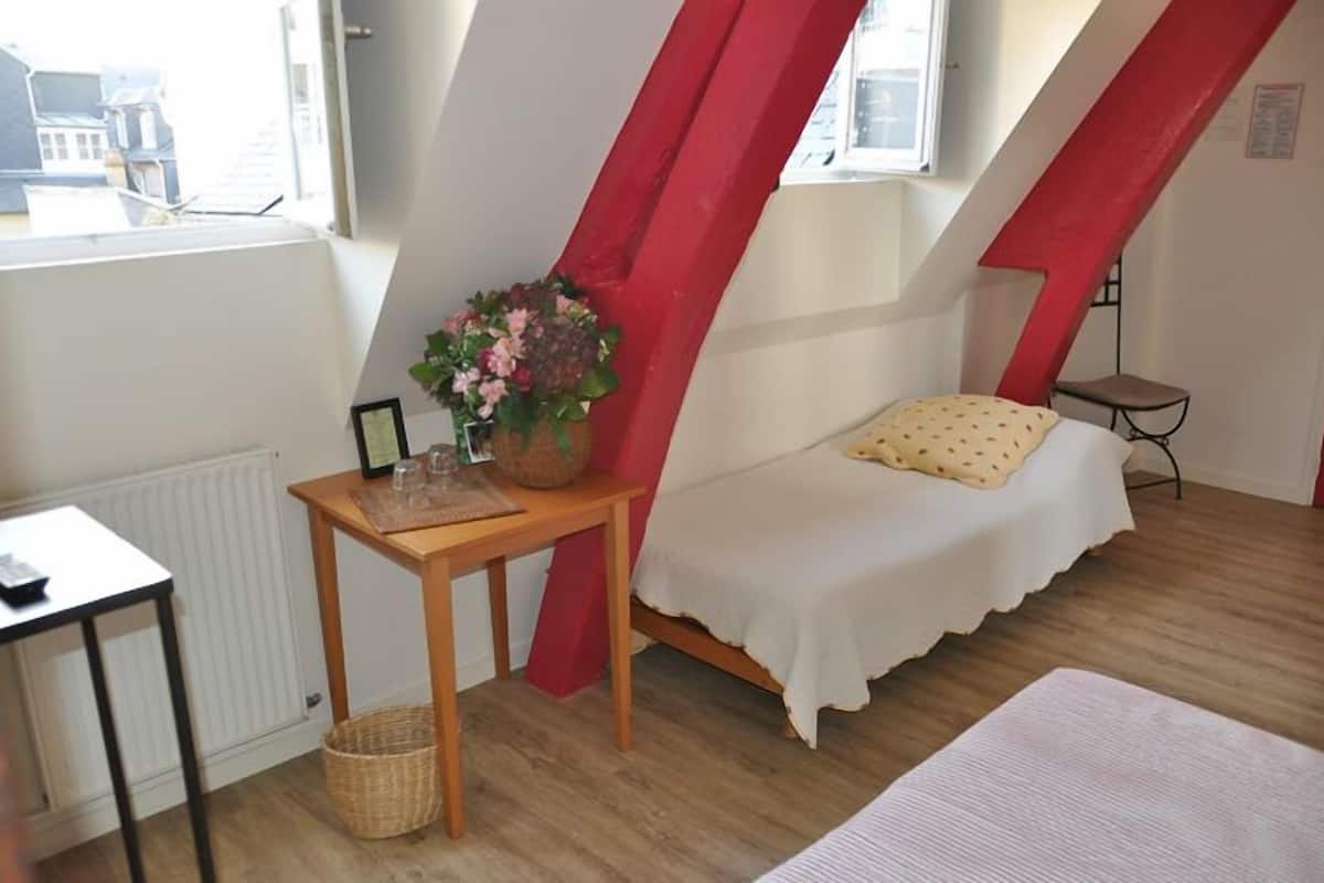 Une petite pièce agréable et chaleureuse, avec un lit, une table ornée d’un vase de fleurs et une fenêtre donnant sur des immeubles.