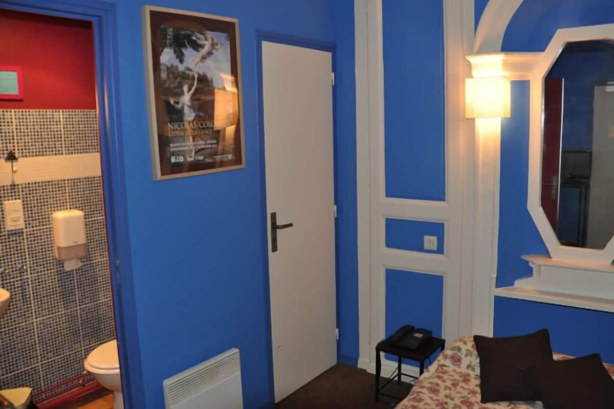 Une chambre avec un mur bleu, une porte blanche, un tableau encadré, une table de chevet, un lit avec une literie à motifs floraux et un miroir.