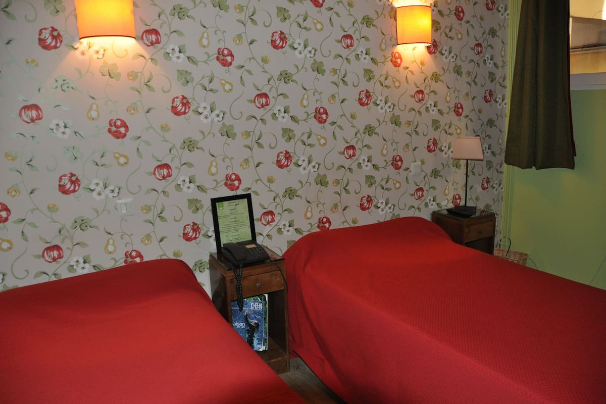 Une chambre d’hôtel avec deux lits, un papier peint à motifs floraux, des lampes de chevet, une petite table avec une lampe et un tableau encadré au mur.