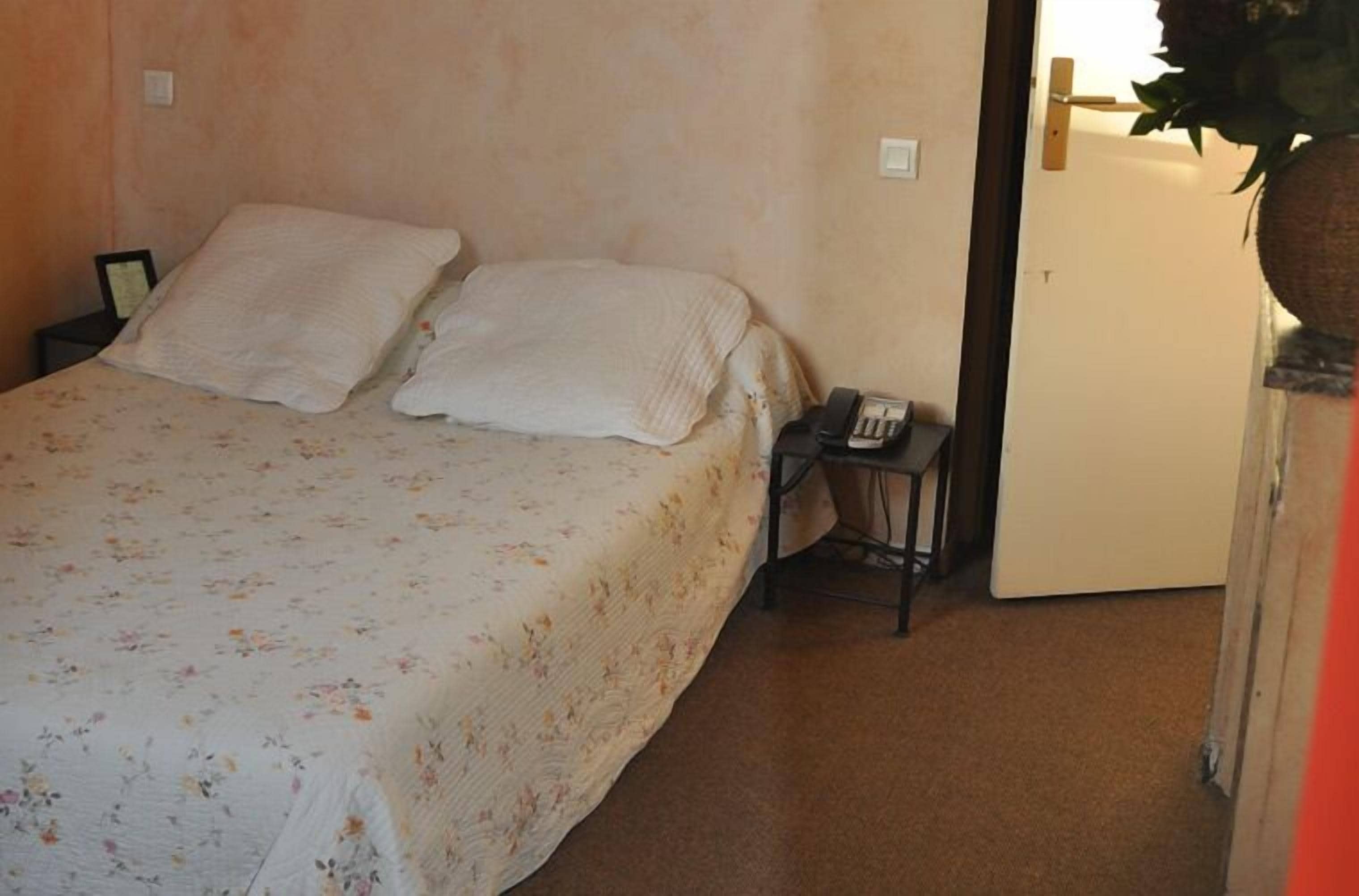 Un lit simple avec une couvre-lit à motifs floraux, une table de chevet avec un téléphone, et une porte ornée d’une plante suspendue.