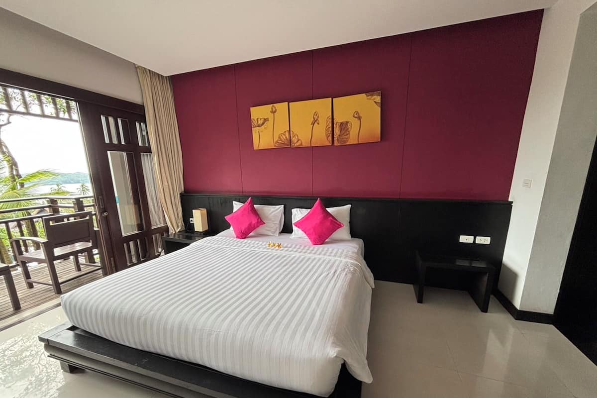 A hotel room with a large bed, a balcony with a view, and a red accent wall.