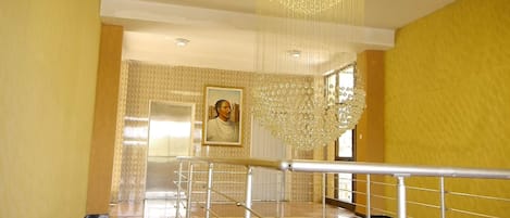 Una escalera con barandilla de metal que lleva a un rellano. Un candelabro cuelga del techo, y se ve un retrato enmarcado en la pared.