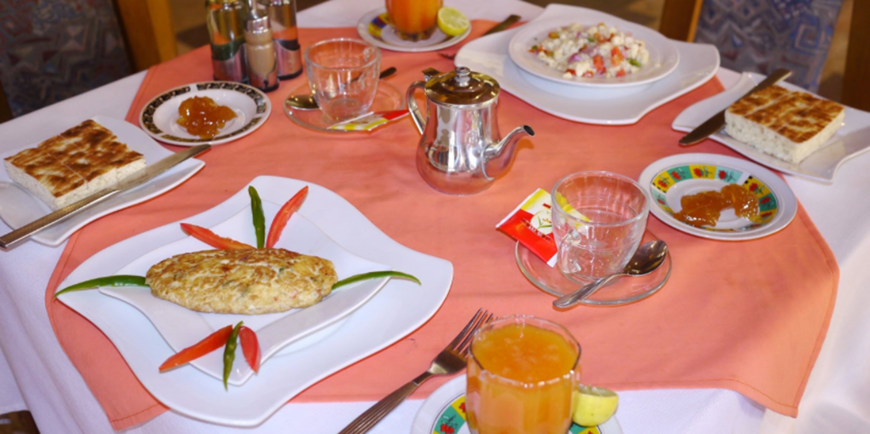 Uma mesa posta com diversos pratos, incluindo um omelete, uma salada de frutas e uma fatia de bolo, além de xícaras e um bule.