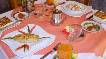 Uma mesa posta com diversos pratos, incluindo um omelete, uma salada de frutas e uma fatia de bolo, além de xícaras e um bule.