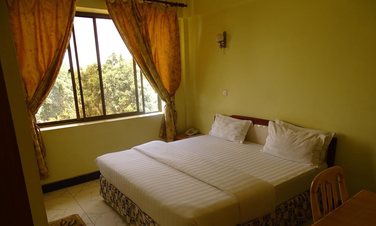 A bedroom with a bed, curtains, a nightstand, and a window with a view of trees.