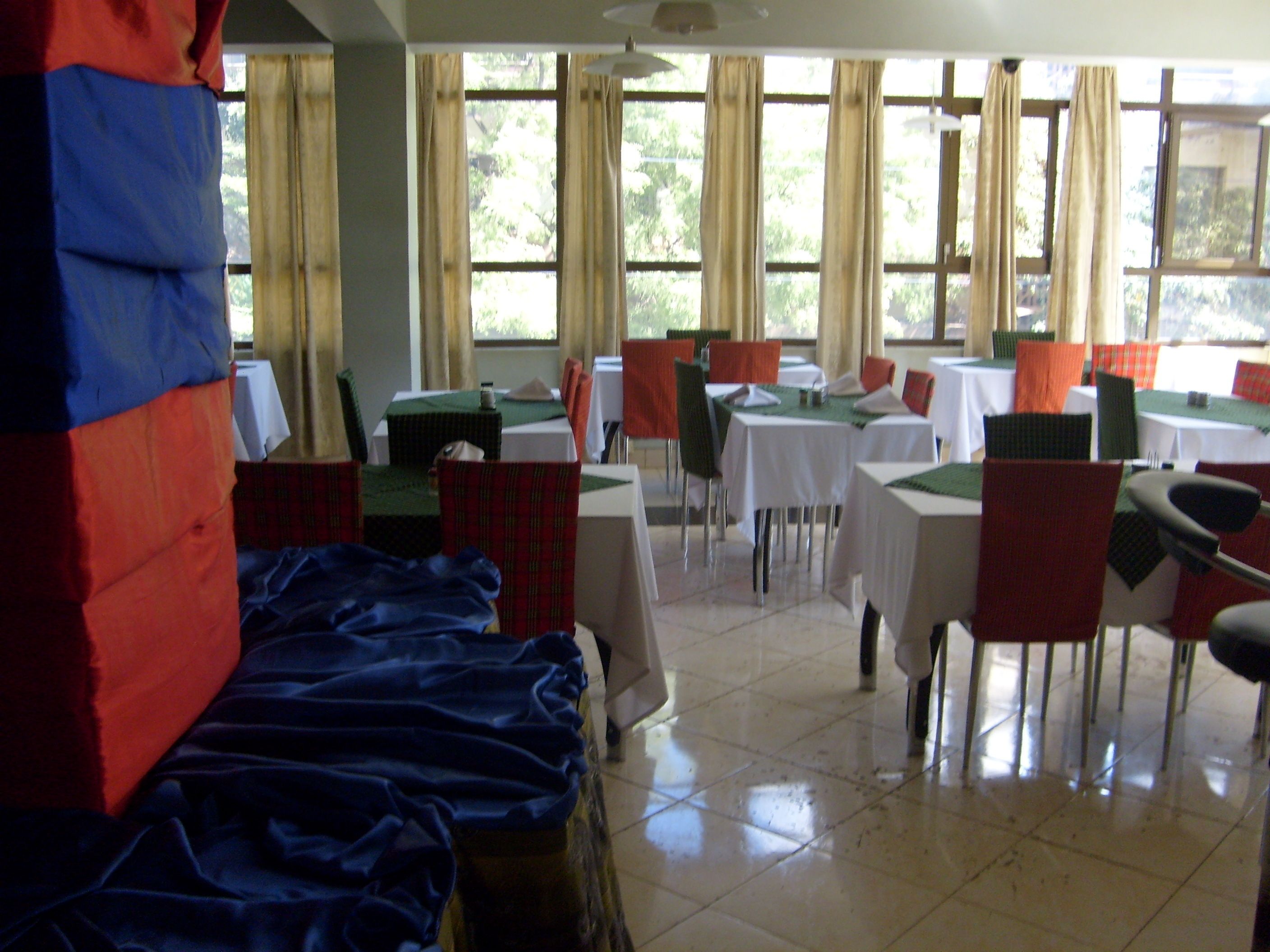 A dining area with tables and chairs, large windows with curtains, and a tiled floor.