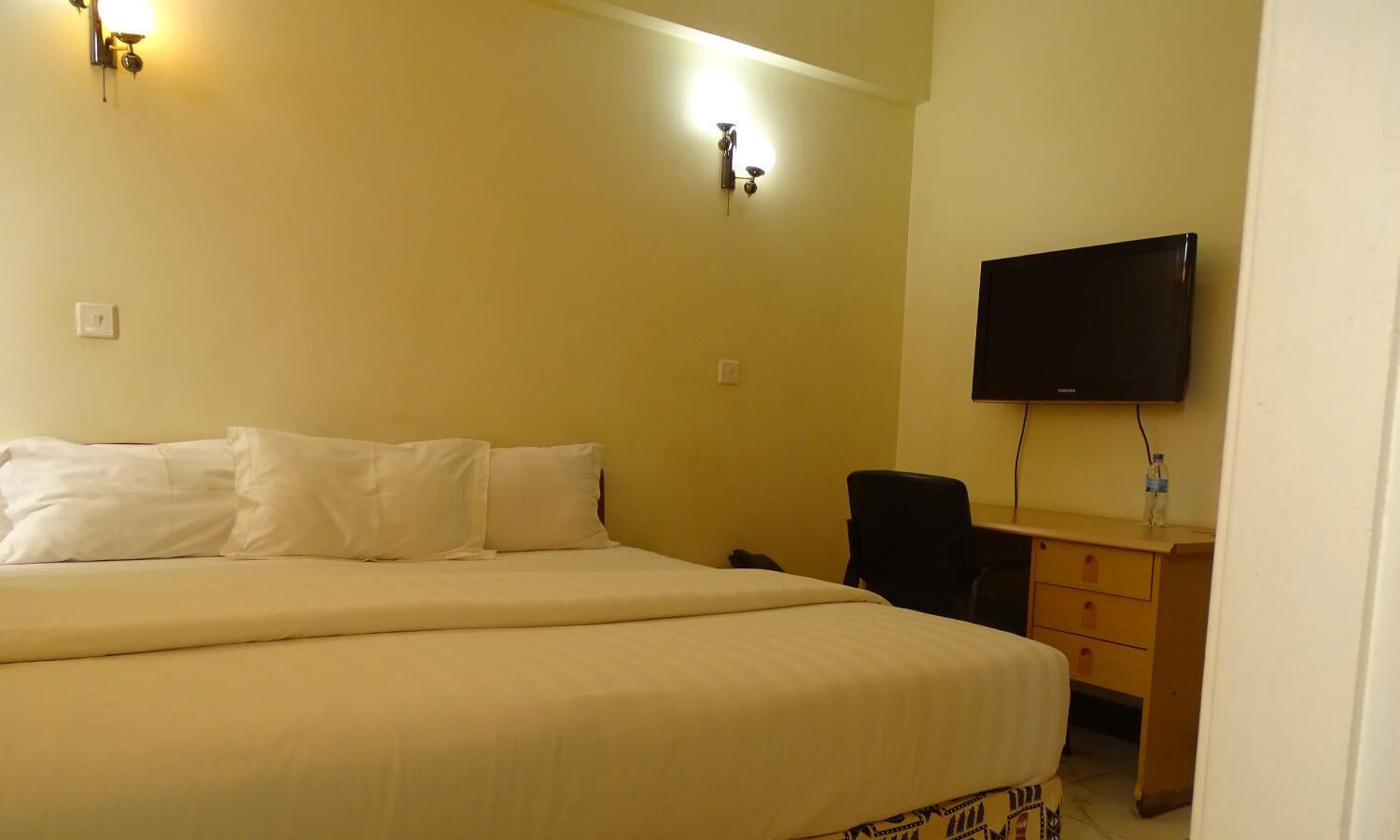 A hotel room with a large bed, a desk with a chair, a flat-screen TV, and wall-mounted lighting.